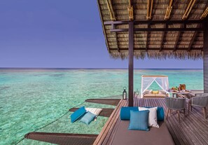 A wooden deck with a thatched roof, a cushioned seating area with pillows, and a table set for dining, overlooking a clear turquoise sea.