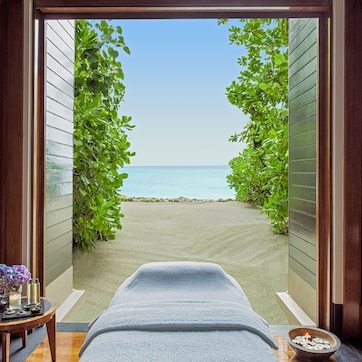A room with a massage table, a small table with candles and flowers, and a view of a beach through an open doorway.