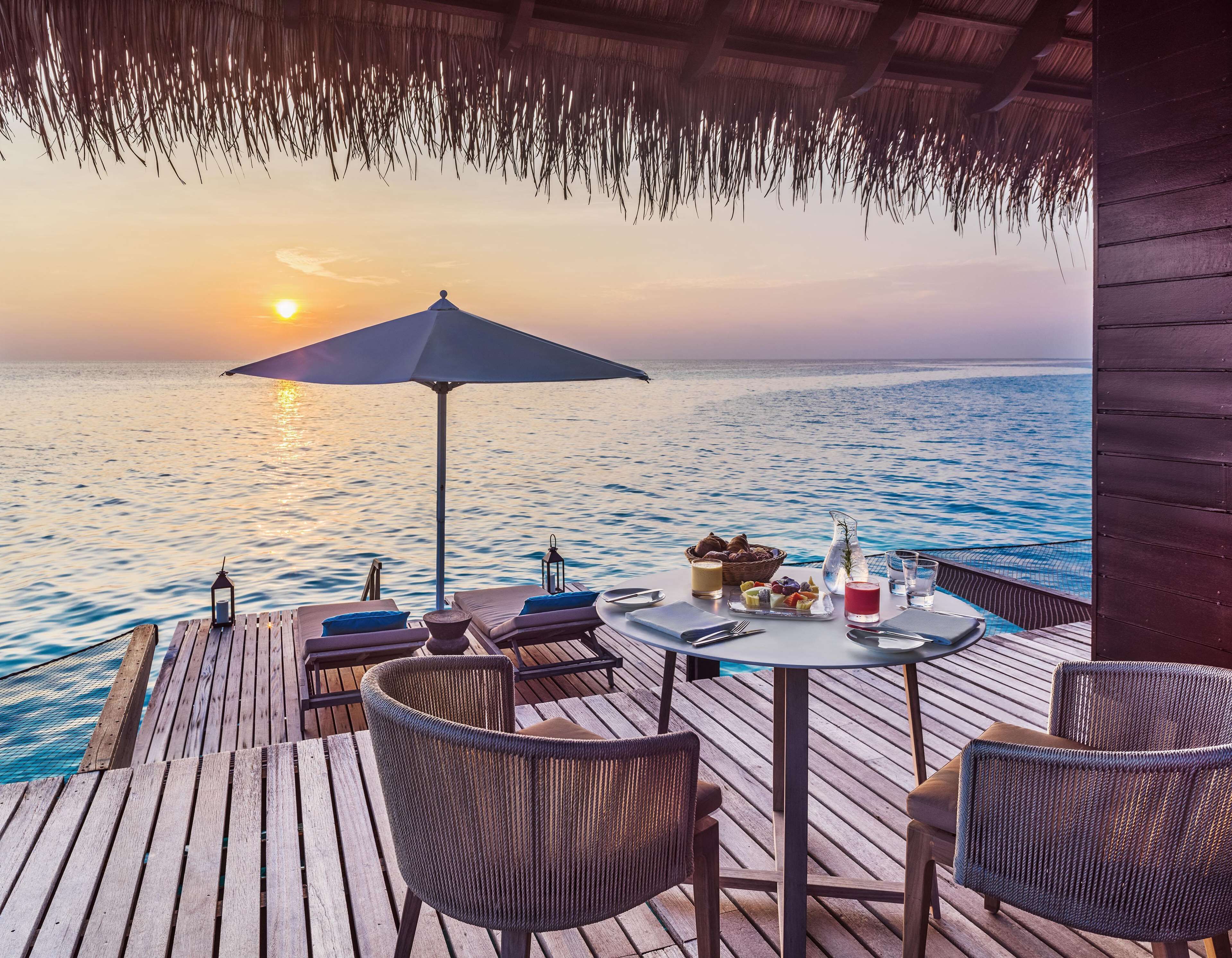 Una terraza de madera con sillas de mimbre y una mesa lista para el desayuno, con vista al mar tranquilo al atardecer.