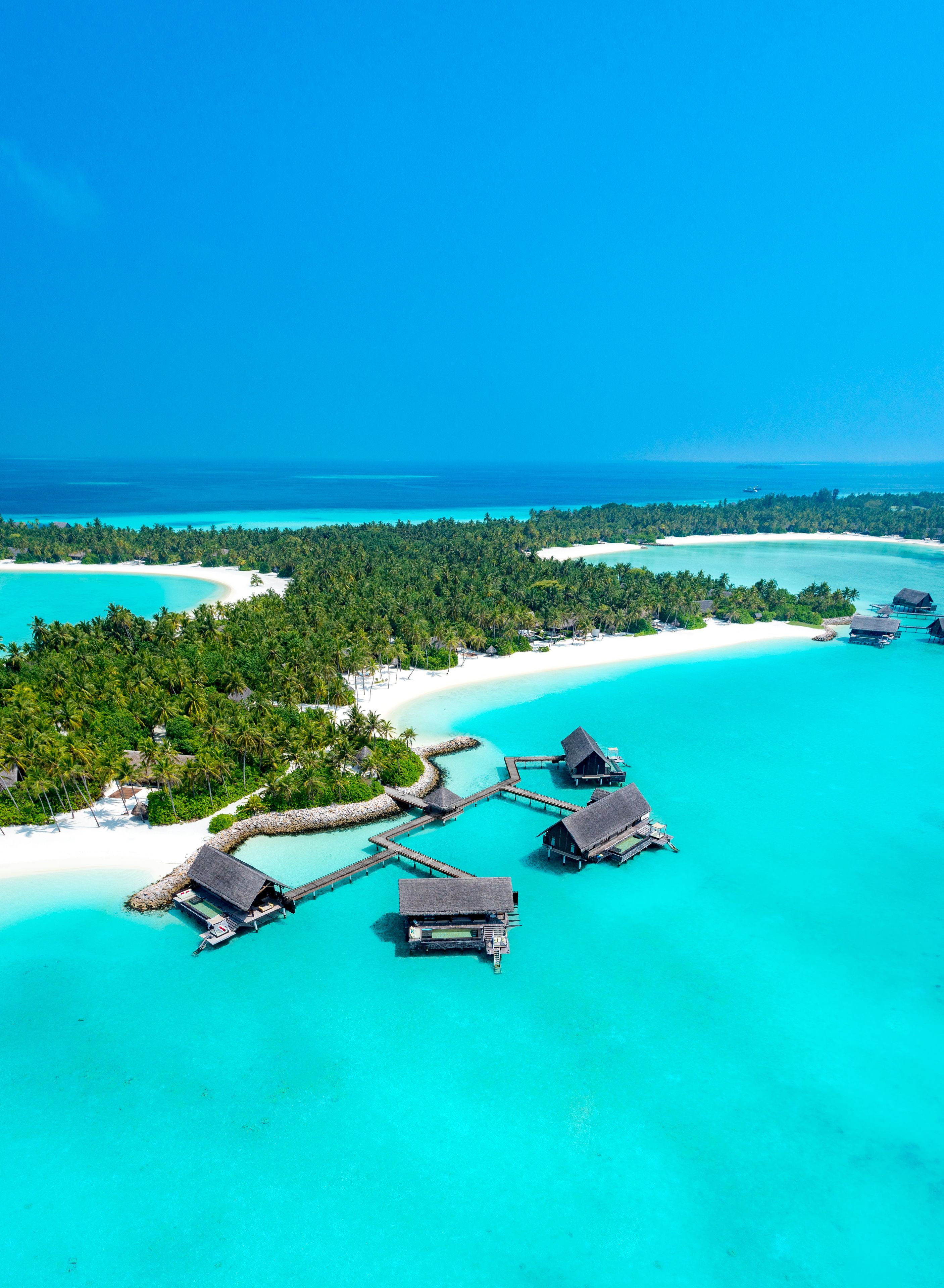Vista aérea de un resort tropical con bungalós sobre el agua, conectados por pasarelas, rodeado de aguas turquesas y una playa de arena blanca.