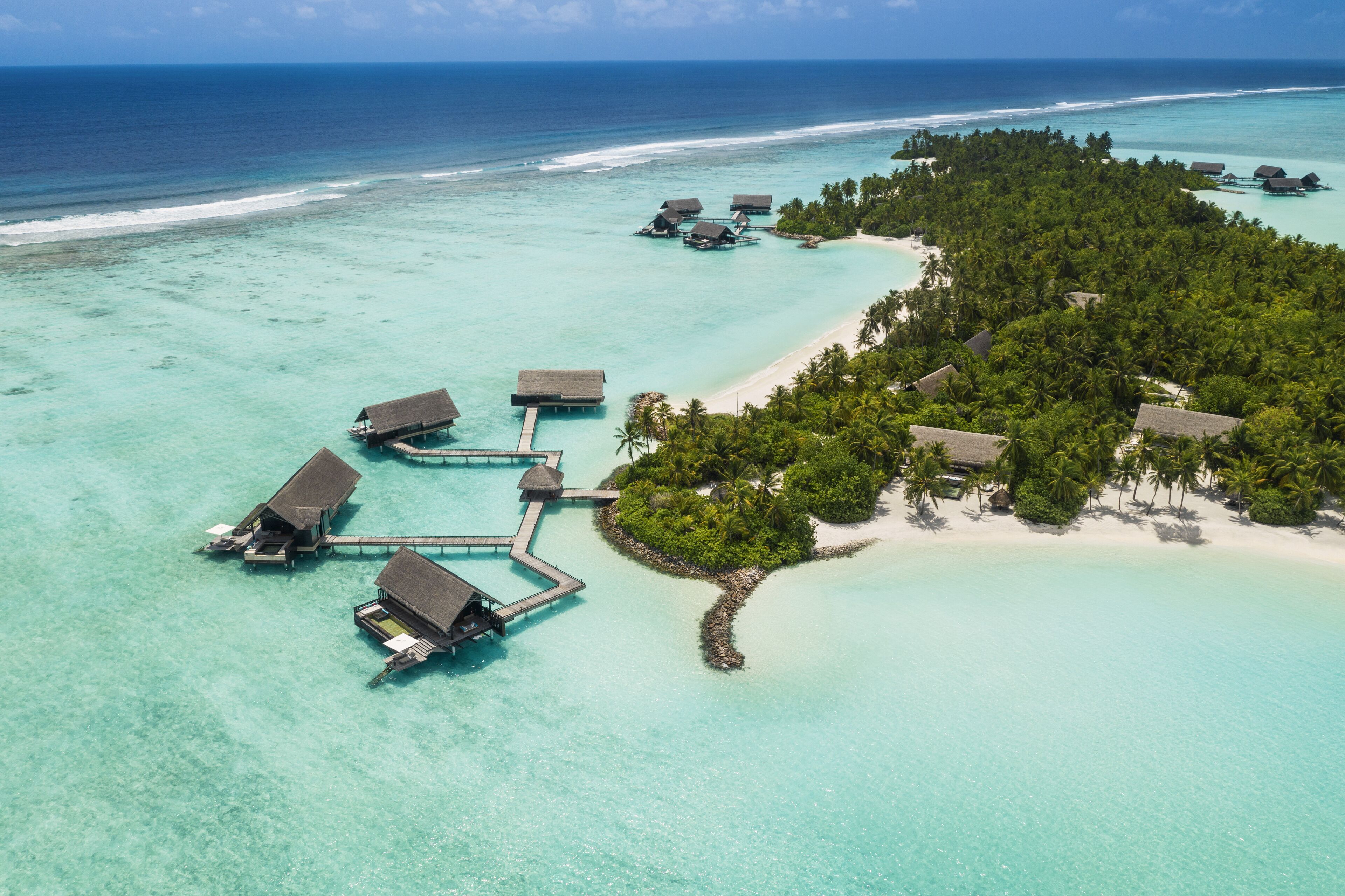 Vista aérea de un resort sobre el agua con varias cabañas conectadas por pasarelas, rodeado de aguas turquesas cristalinas y una pequeña isla con vegetación exuberante.