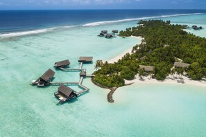 An aerial view of an overwater resort with multiple bungalow-style accommodations connected by walkways, surrounded by clear turquoise waters and a small island with dense vegetation.