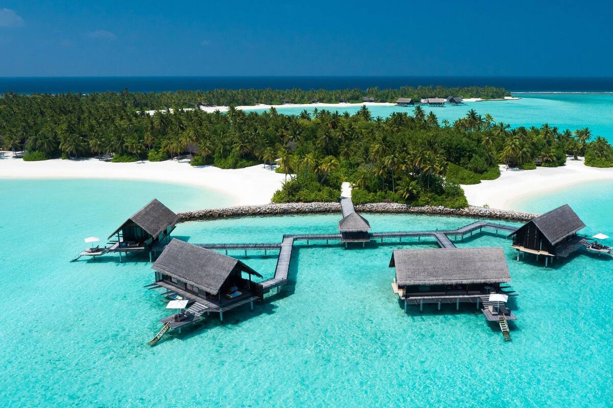 Une vue aérienne de bungalows sur pilotis reliés par une passerelle en bois au-dessus d’eaux turquoise, entourés d’une végétation luxuriante et de plages de sable blanc.