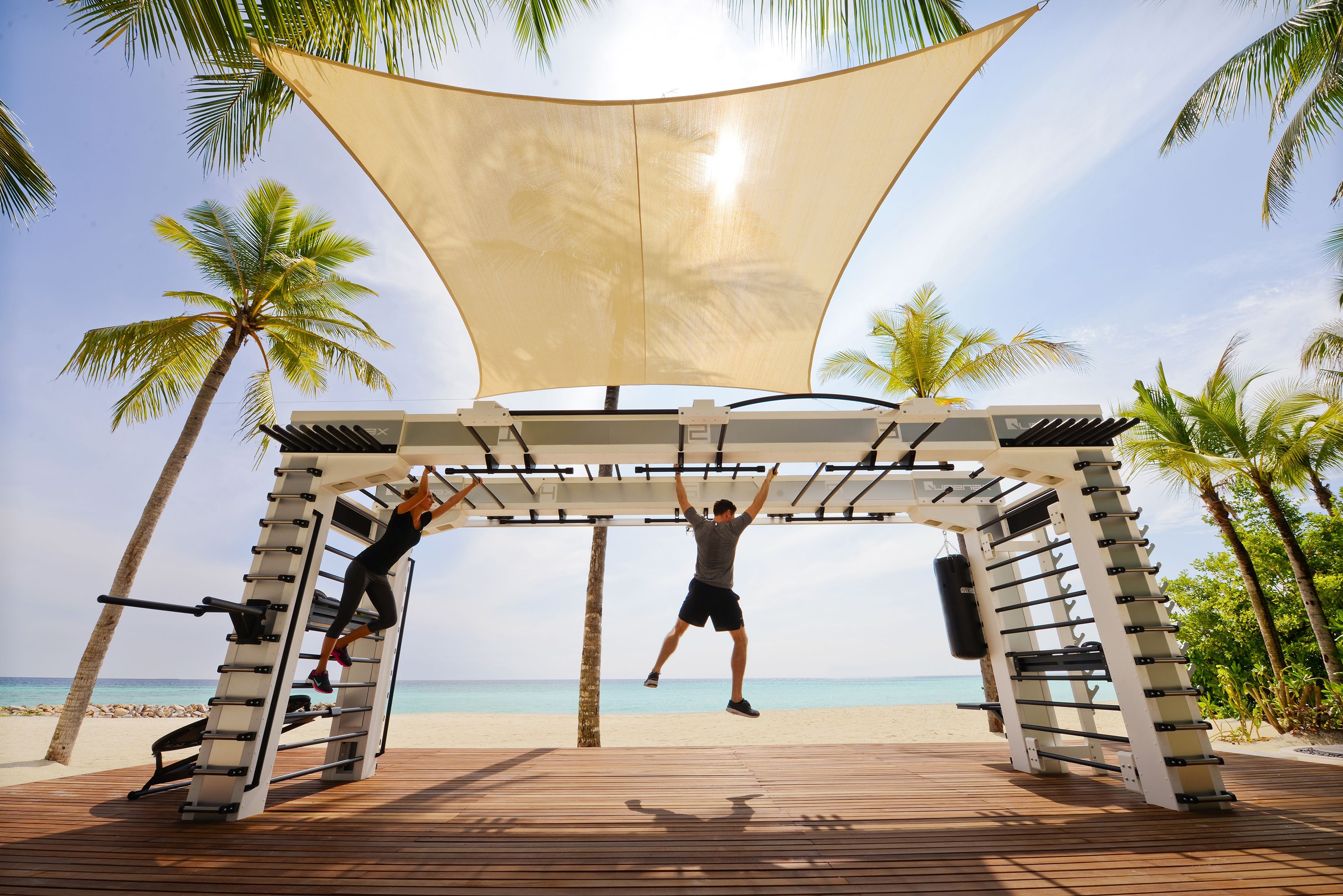 Zona de gimnasio con barra de dominadas y suelo de madera, con vista a una playa con palmeras.