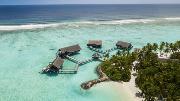 An aerial view of overwater bungalows connected by walkways, surrounded by turquoise waters and palm trees.