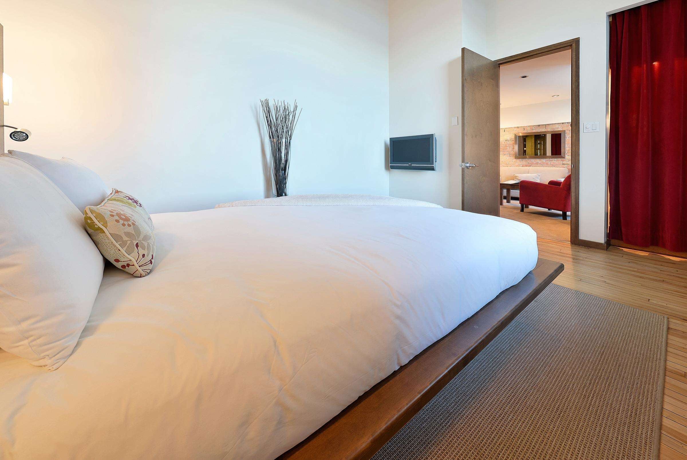 A hotel room with a large bed, a flat-screen TV mounted on the wall, and a view into the living area through an open door.
