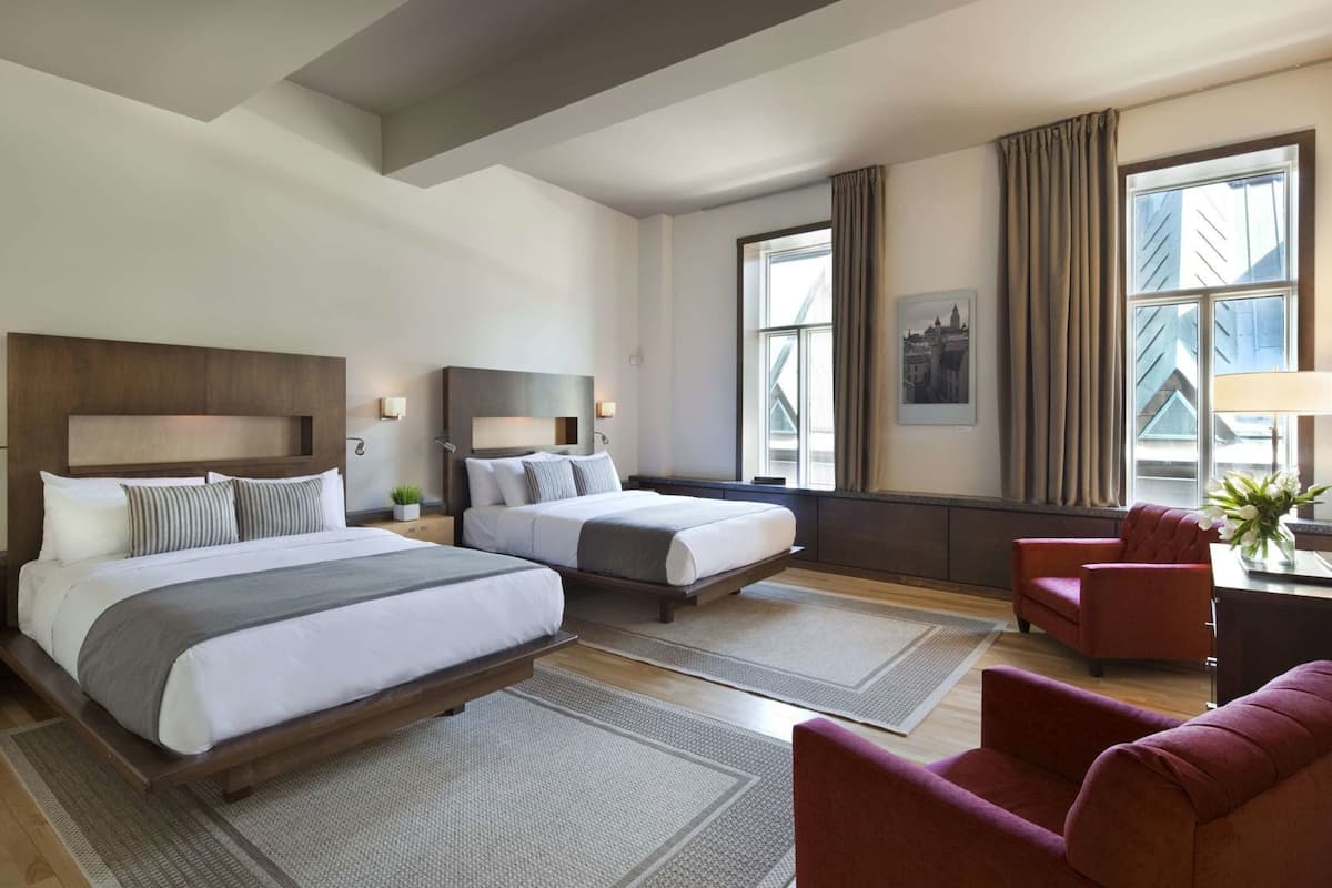 A hotel room with two beds, a red armchair, a desk with a lamp, and large windows with curtains.