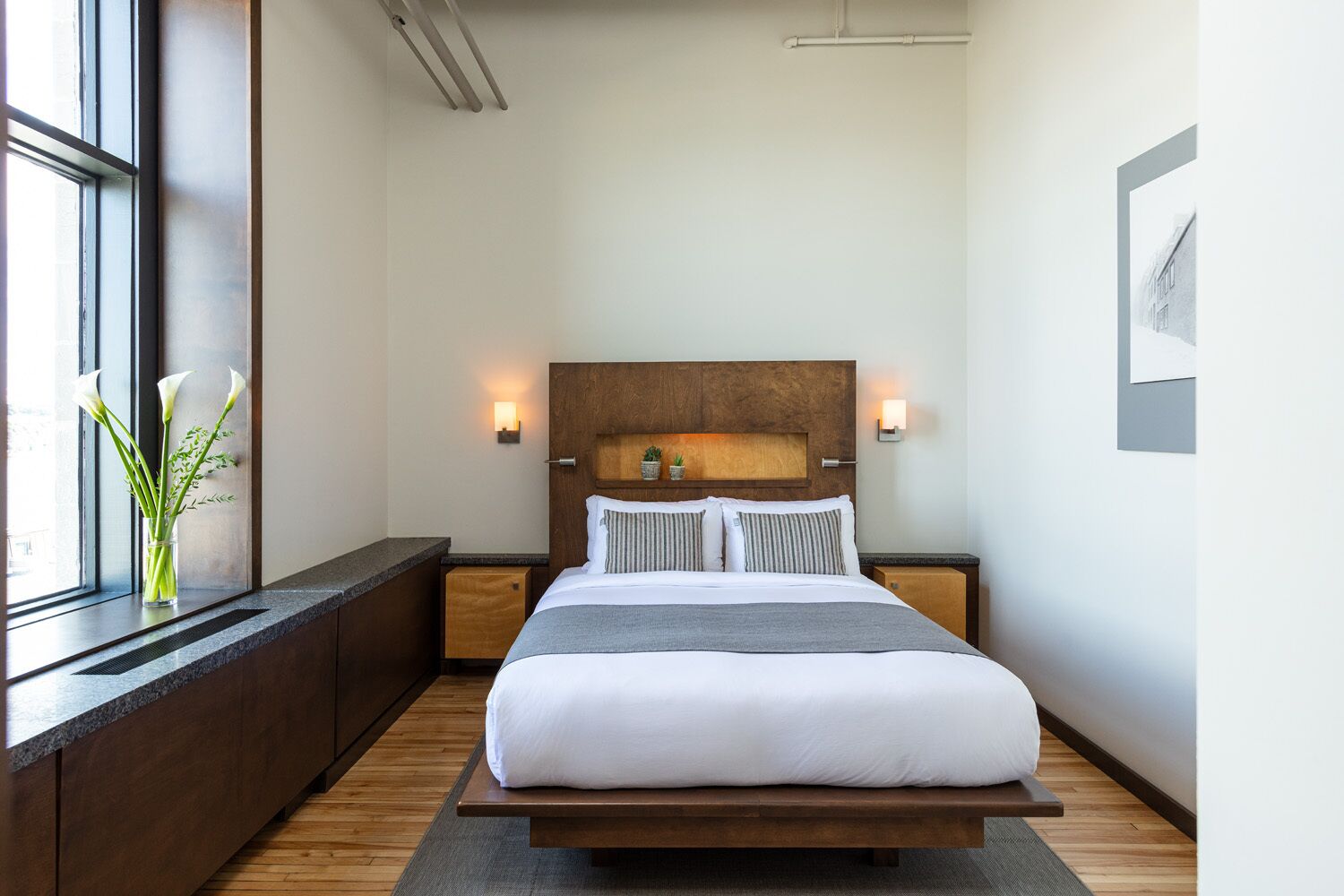 Une chambre moderne avec un grand lit, une tête de lit en bois, des tables de chevet, une fenêtre avec vue et un vase rempli de fleurs.