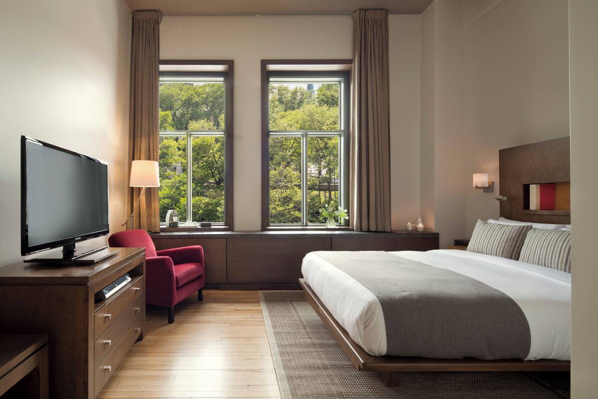 A modern hotel room with a large bed, a TV on a wooden dresser, a red chair, and a view of trees through two windows.