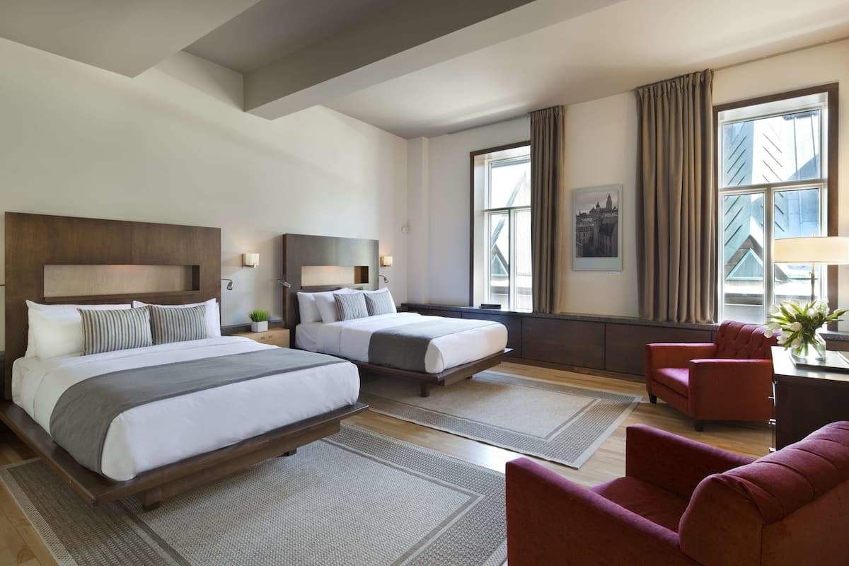 A hotel room with two beds, a desk, a chair, and large windows.