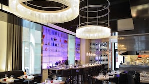 Un restaurant doté de nombreuses tables et chaises, d’un bar avec des étagères remplies de bouteilles et d’un lustre moderne.