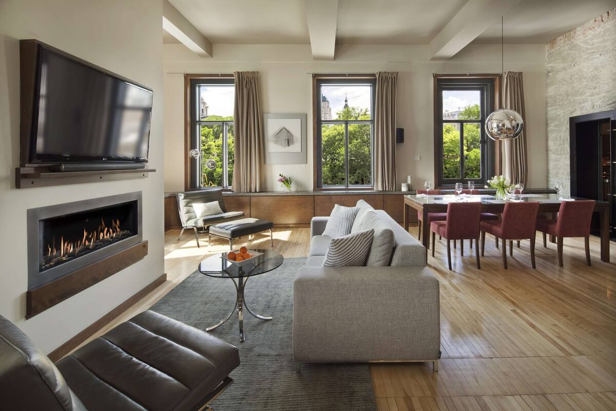 A modern living room with a fireplace, a flat-screen TV, a dining area with red chairs, and large windows with curtains.