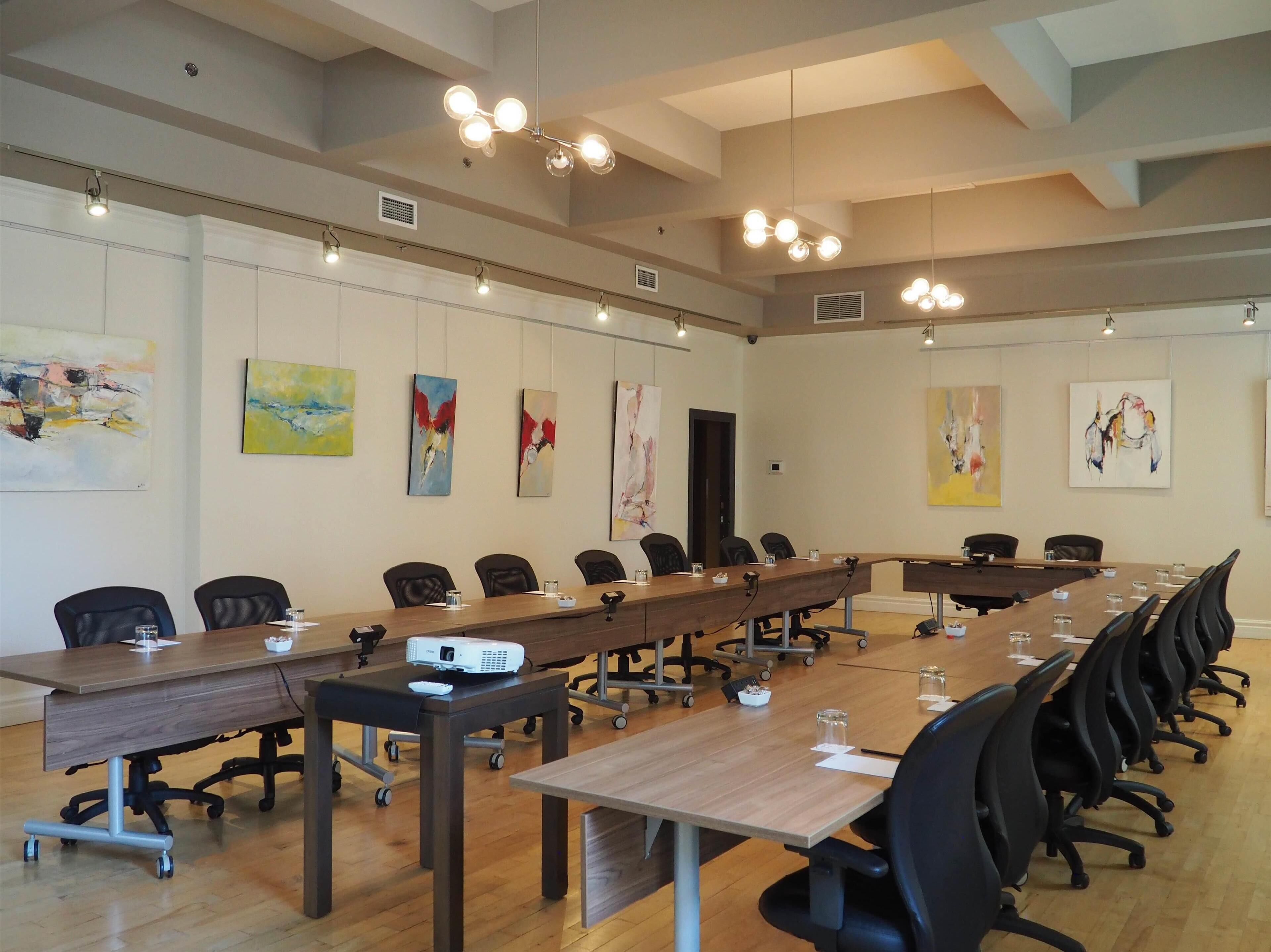 Een congreszaal met een lange tafel, meerdere stoelen, een projectiescherm en kunstwerken aan de wanden.