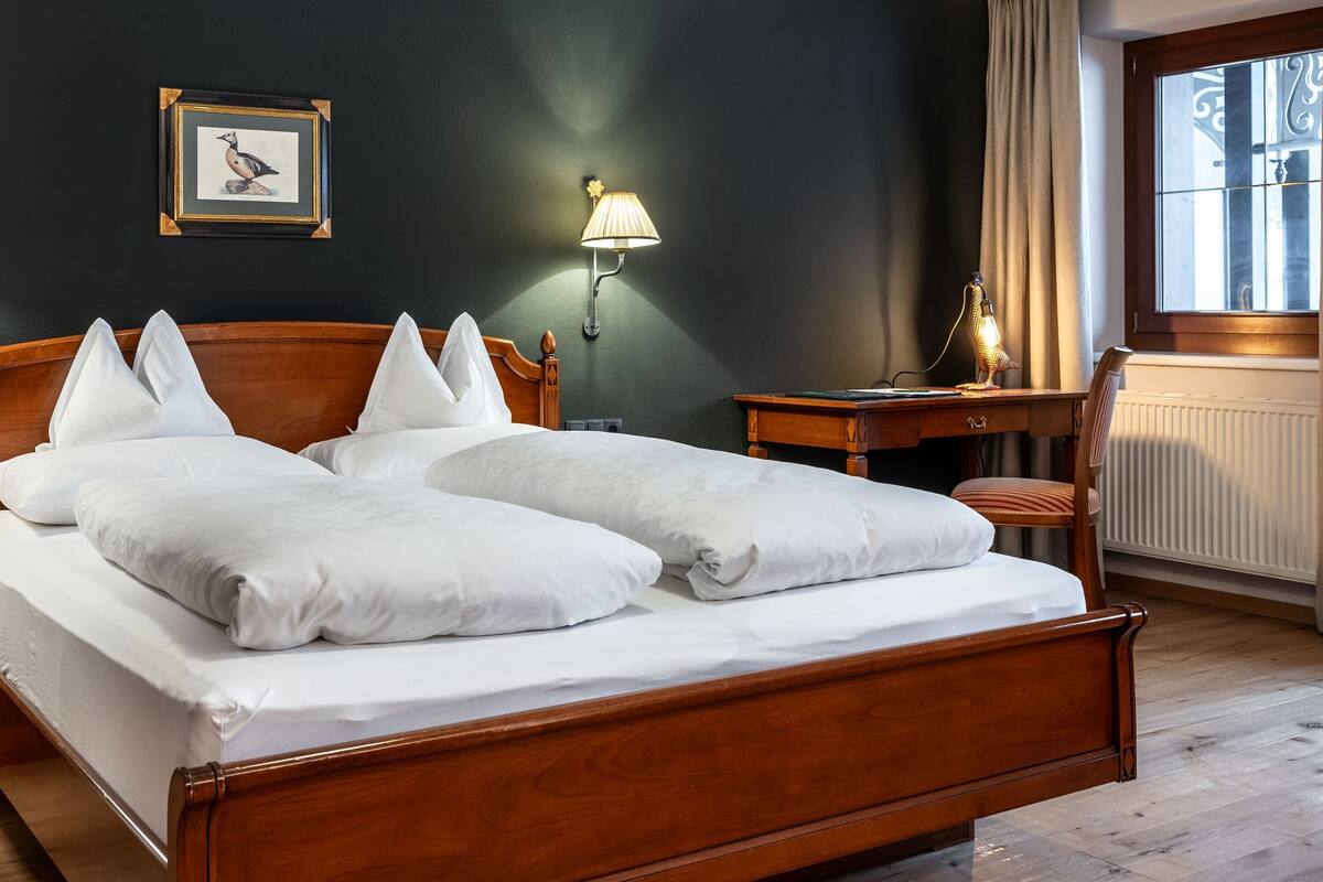 Una cama bien hecha con sábanas y almohadas blancas, un cabecero de madera, dos lámparas de noche, un cuadro en la pared y una ventana con cortinas.