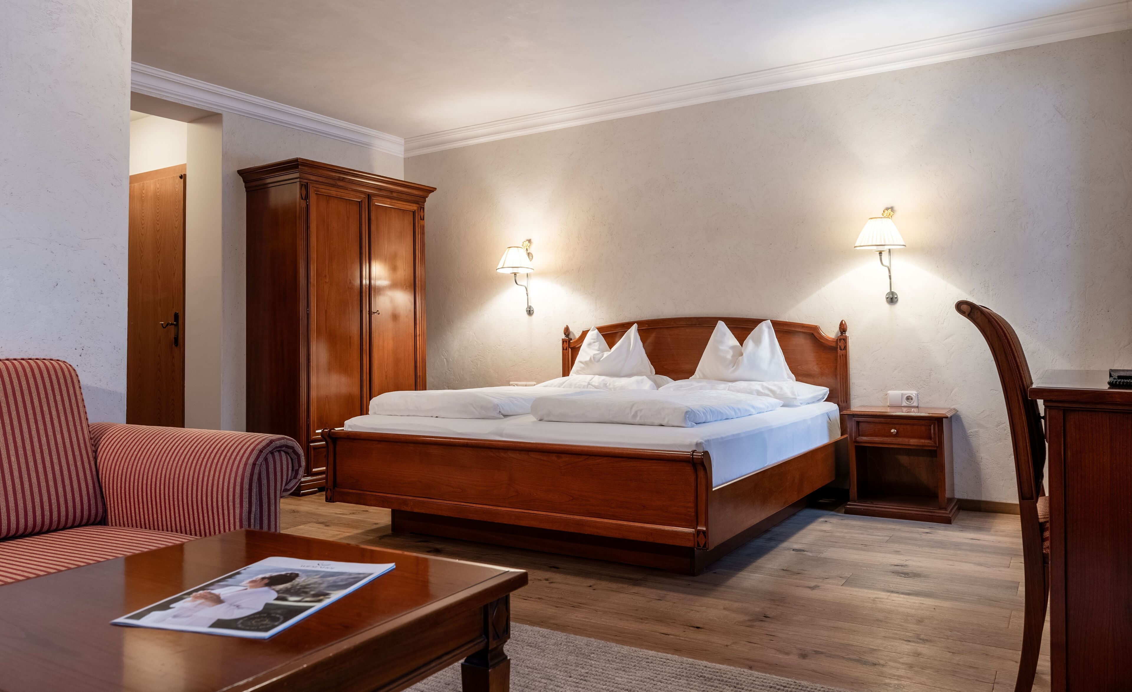 Quarto de hotel com cama de madeira, guarda-roupa de madeira, mesa de cabeceira, luminária, cadeira e revista sobre uma mesa de centro.