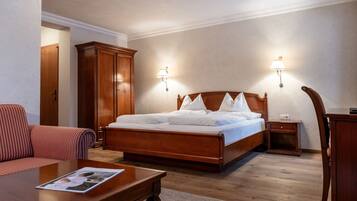 Quarto de hotel com cama de madeira, guarda-roupa de madeira, mesa de cabeceira, luminária, cadeira e revista sobre uma mesa de centro.