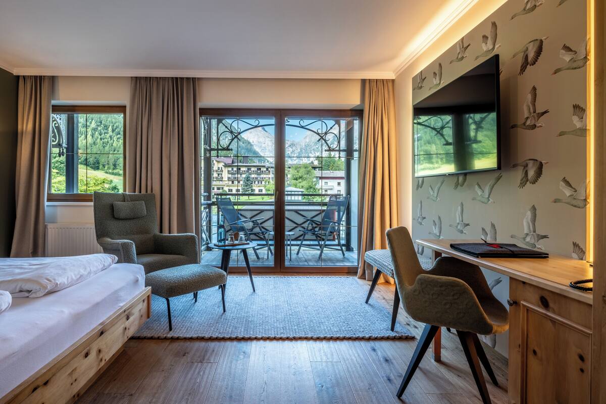 Habitación de hotel con cama, escritorio, silla y balcón con vistas a montañas y edificios.