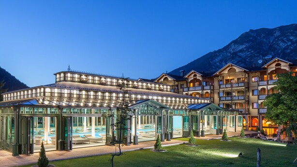 Un hotel con zona de piscina cubierta de vidrio, varios balcones y un telón de fondo montañoso.