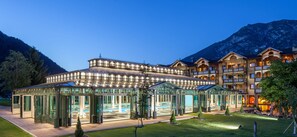 Um hotel com área de piscina envidraçada, várias varandas e montanhas ao fundo.