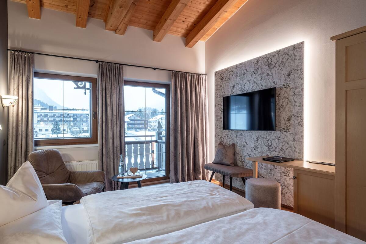 Habitación de hotel con cama, dos sillones, una mesita, televisor y ventana con vista a edificios y montañas.