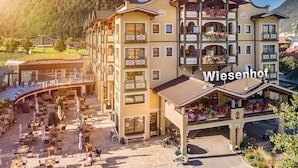Un hotel de varios pisos con balcones, mobiliario exterior y montañas al fondo.