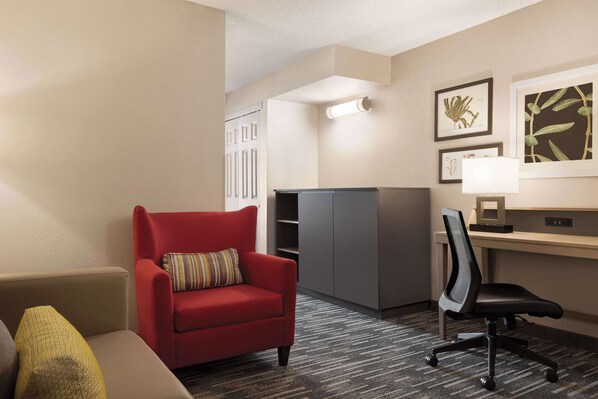 A hotel room with a red armchair, a desk with a computer, a lamp, and framed botanical artwork on the wall.