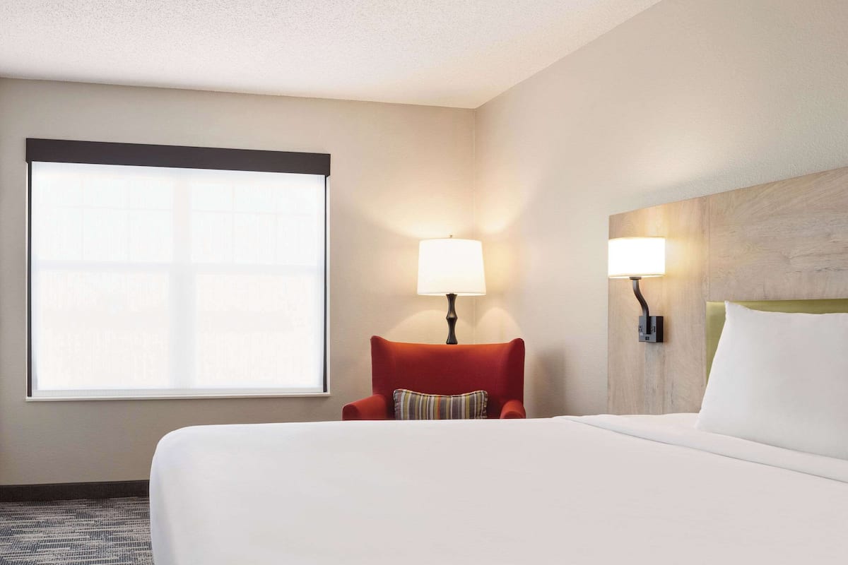 A hotel room with a large bed, a red armchair, two wall-mounted lamps, and a large window with blinds.
