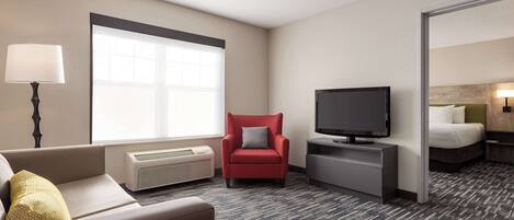 A hotel room with a sofa, a red armchair, a TV, a bed, and a large window.
