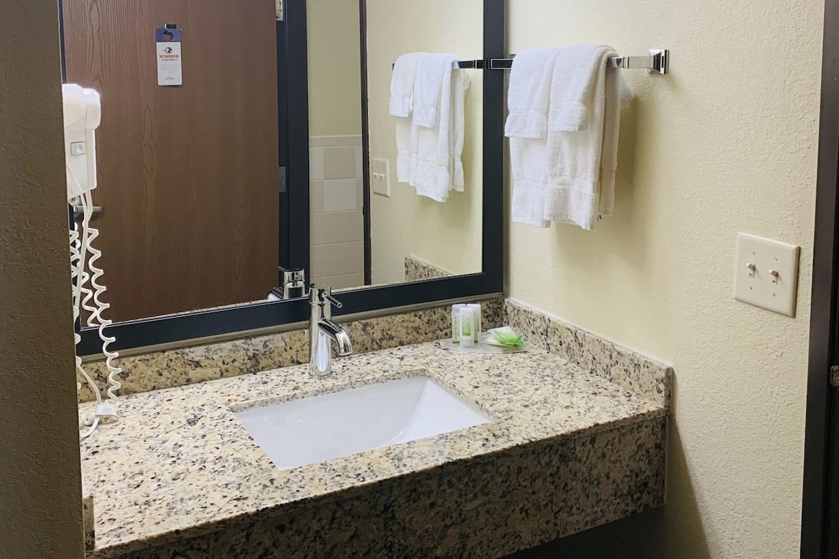 A bathroom with a granite countertop sink, a mirror with lights, and a towel rack with white towels.