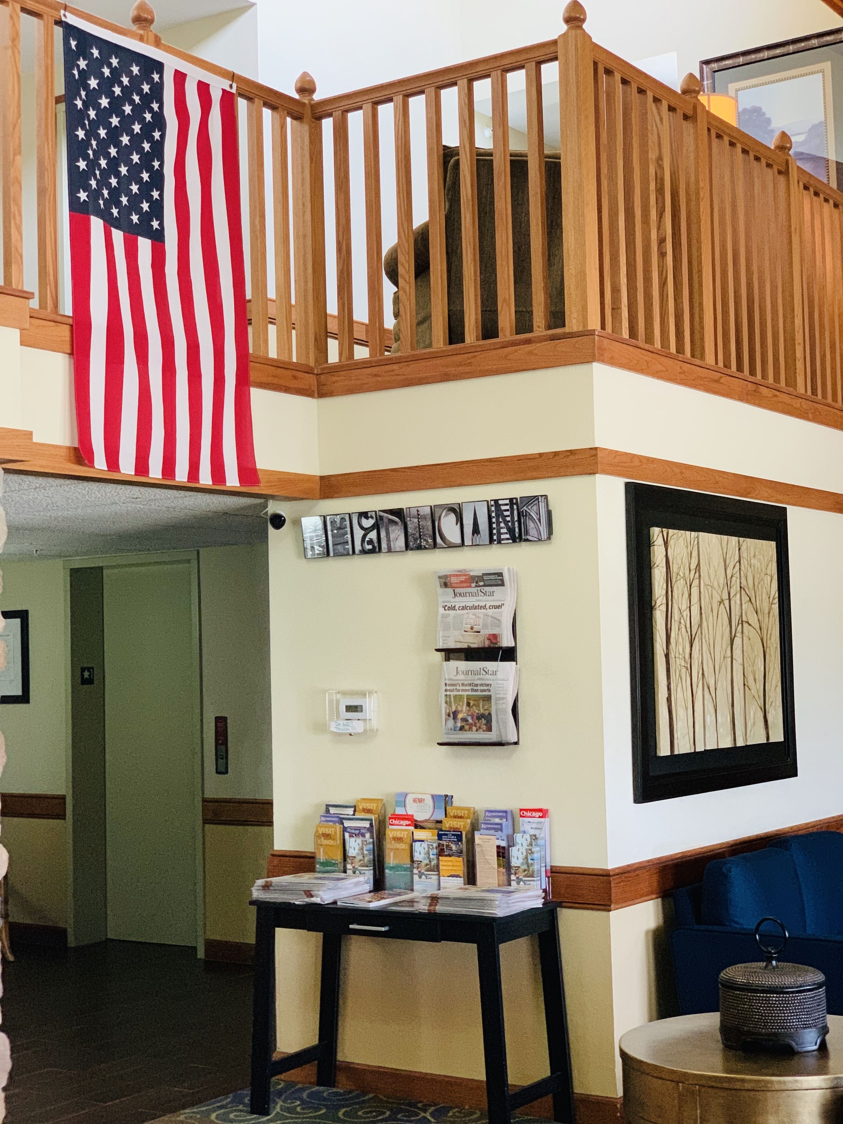 Un hall d’hôtel avec un escalier en bois, un drapeau américain, un présentoir à journaux fixé au mur, un tableau encadré et une table avec des magazines.