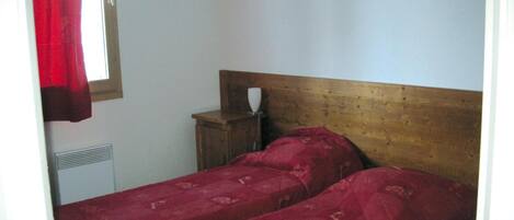 Une petite chambre simple, avec un lit doté d’une tête de lit en bois, une table de chevet et une fenêtre ornée d’un rideau rouge.