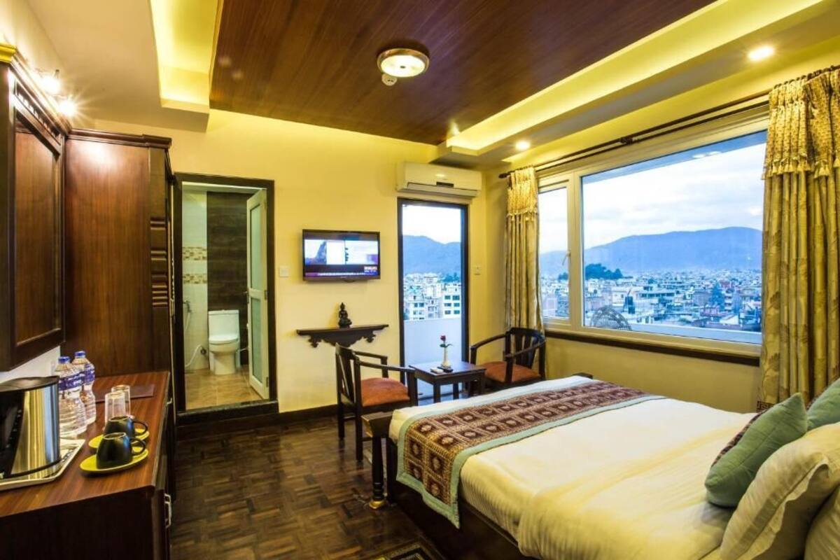 A hotel room with a bed, a desk with a laptop, a TV, and a view of a cityscape through a large window.