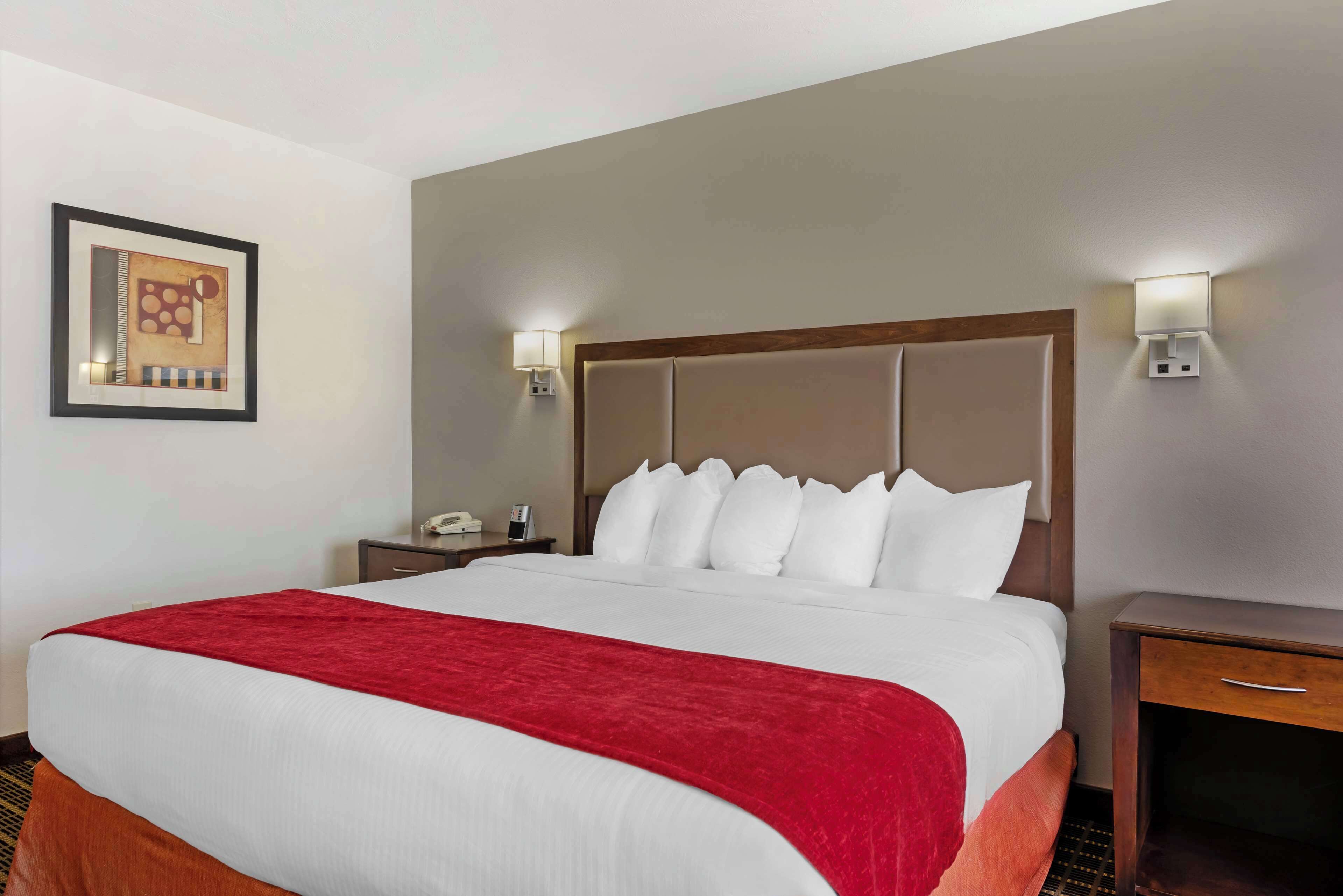 A hotel room with a large bed, a red throw blanket, a wooden headboard, two bedside tables with lamps, and a framed picture on the wall.