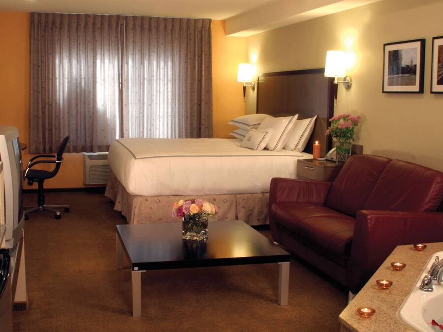 A hotel room with a bed, a desk, a sofa, a bathroom with a sink and mirror, and a dining area with a chair and a table.