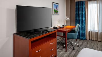 A hotel room with a flat-screen TV, a desk with a lamp, a chair, and a bed.