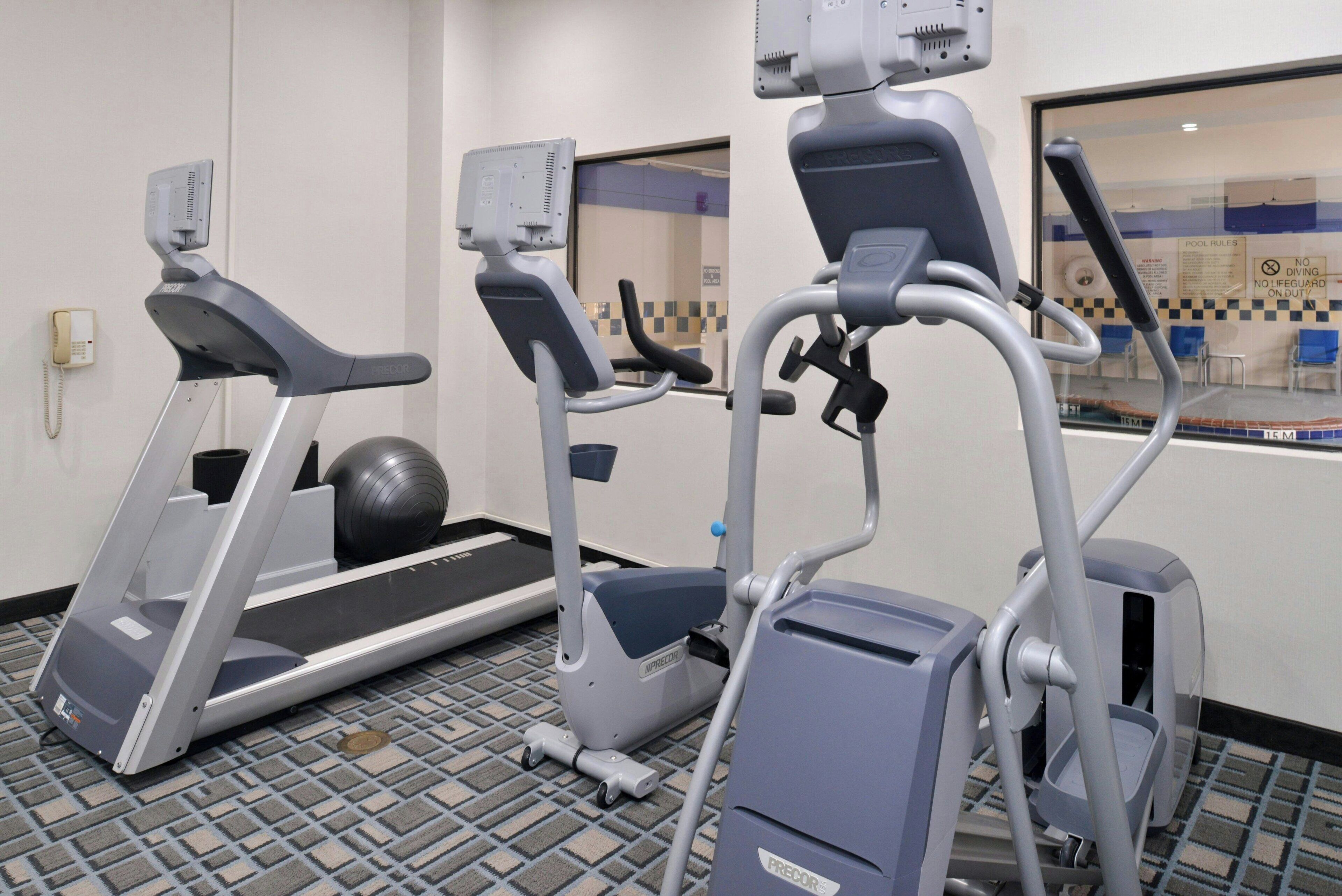Un gimnasio con cintas de correr, una elíptica y una bicicleta estática, con alfombra estampada y una ventana que da a una piscina.