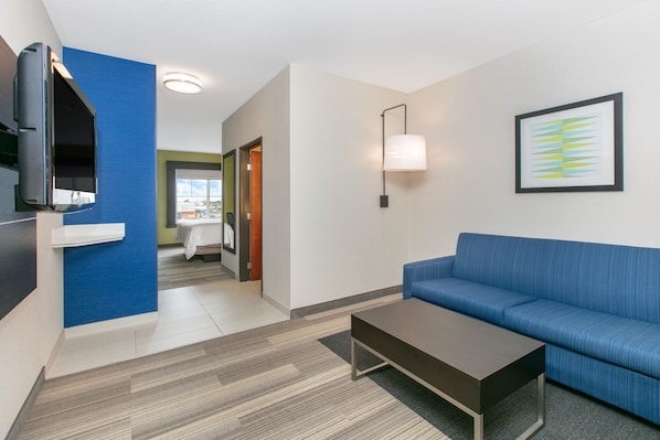 Une chambre d’hôtel moderne avec un canapé bleu, un téléviseur à écran plat, un luminaire fixé au mur et une œuvre d’art abstraite encadrée.
