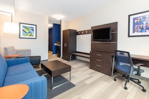 Une chambre d’hôtel moderne avec un canapé bleu, une petite table orange, un bureau avec une chaise, un téléviseur à écran plat et une armoire.