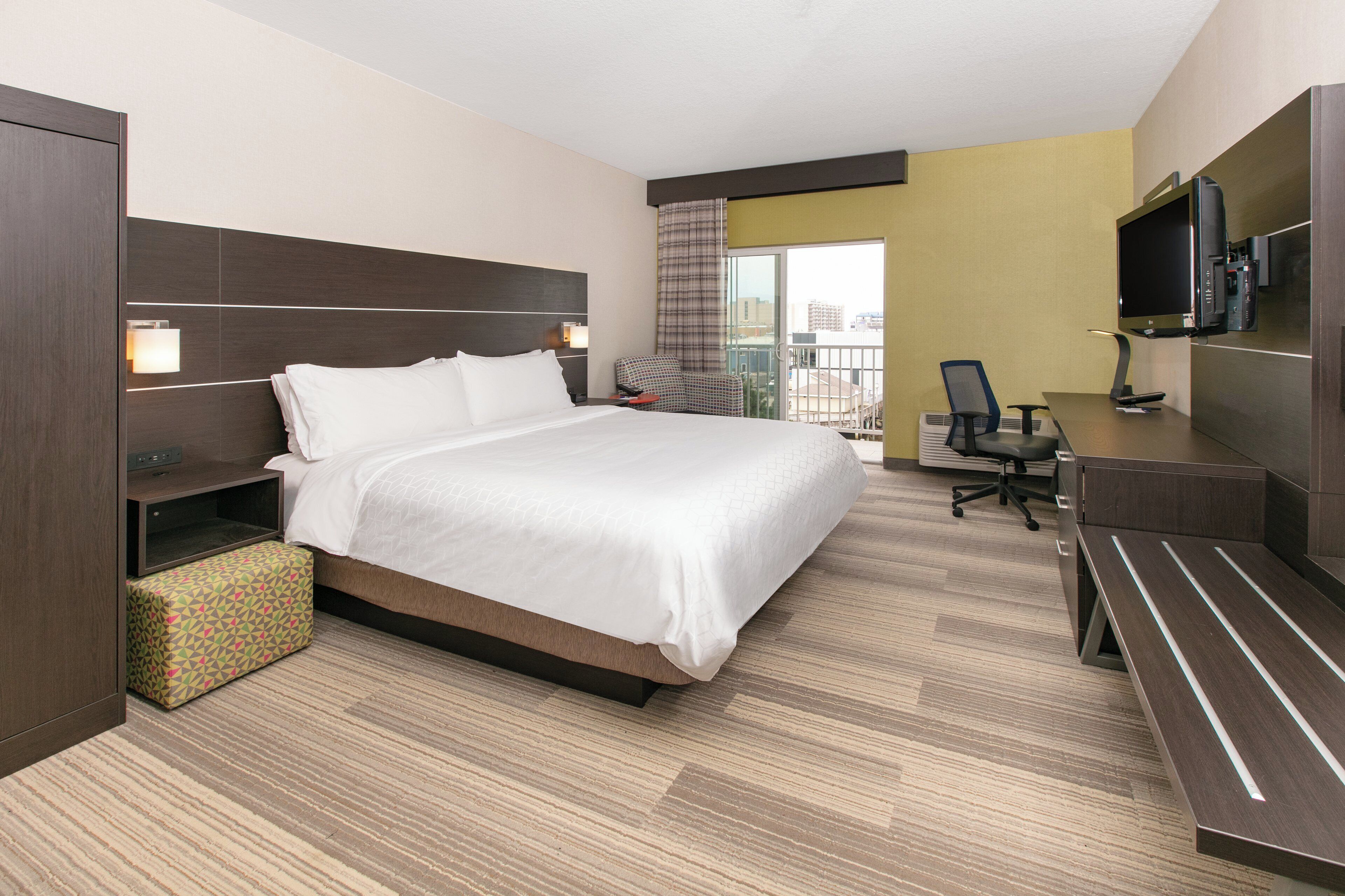 A hotel room with a bed, desk, chair, television, and a view of the city.