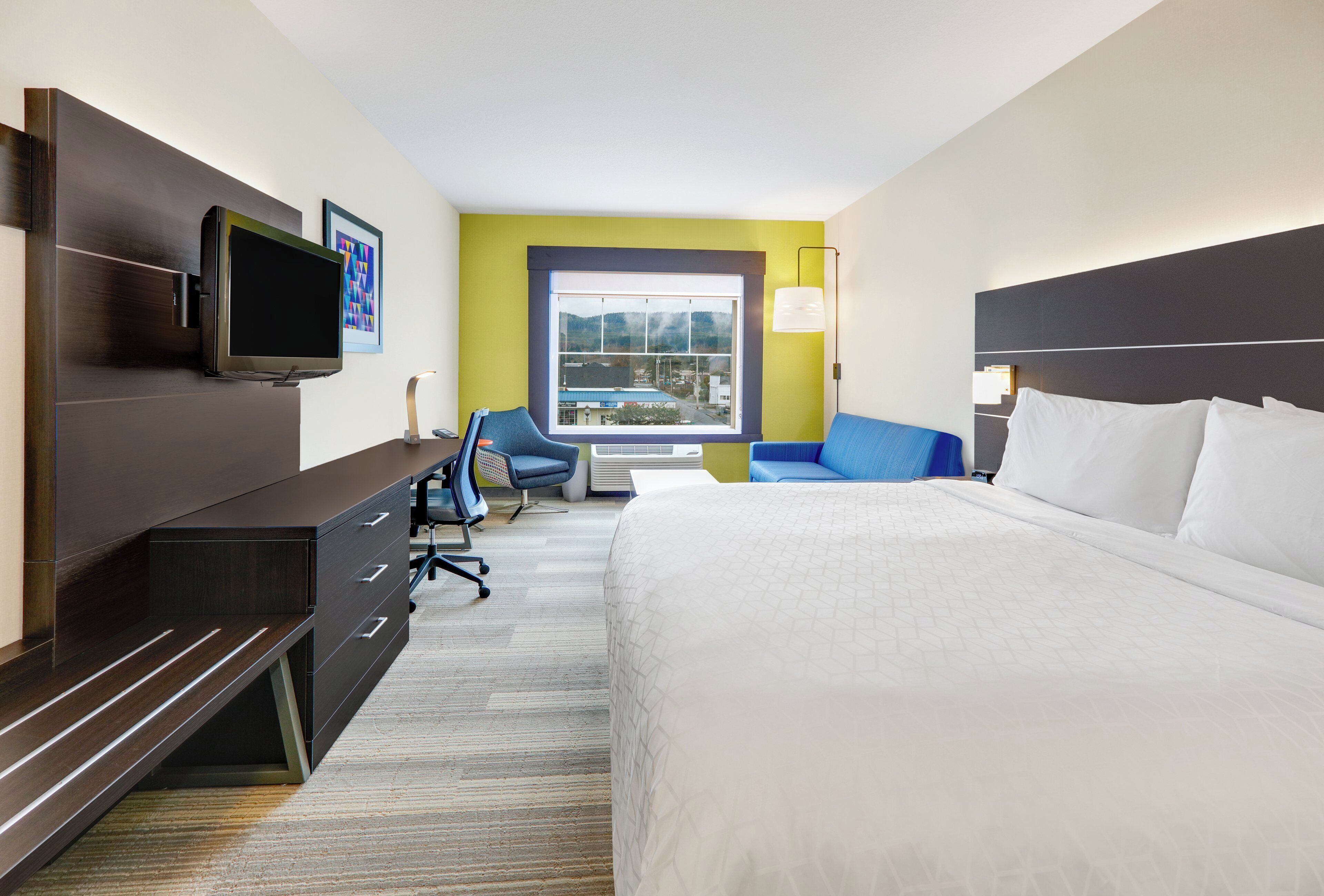 A hotel room with a bed, a TV, a desk, a chair, a window with a view, and a lamp.