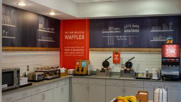 A modern kitchen area with waffle-making equipment, a red sign advertising waffles, and a juice dispenser.