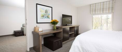 A hotel room with a bed, a desk with a computer, a television, a window with blinds, and a framed picture on the wall.