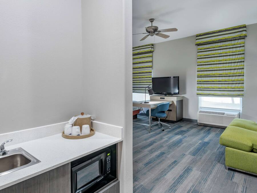 A modern hotel room with a kitchenette, a desk with a computer, a green sofa, and a ceiling fan.