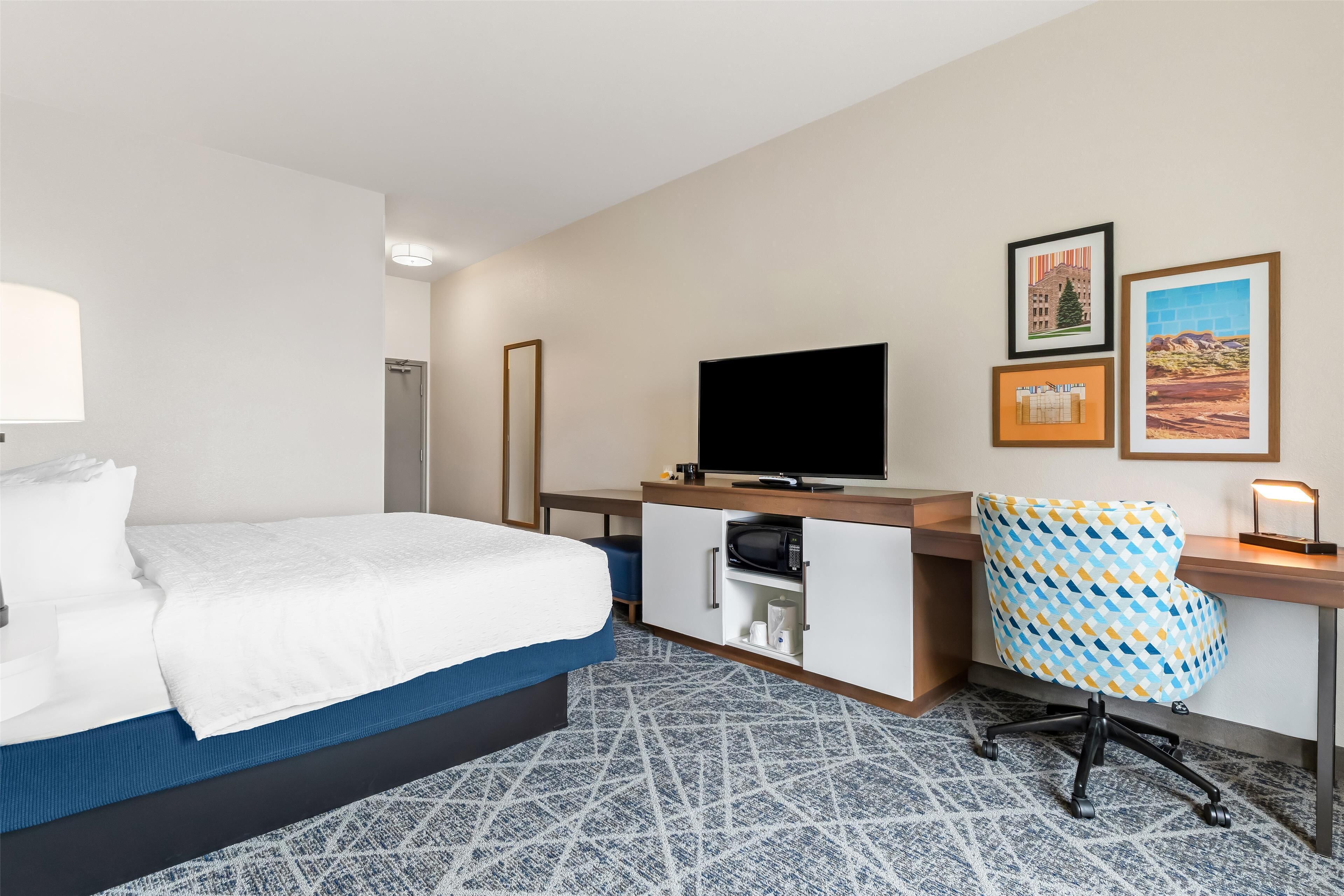 A hotel room with a bed, a desk with a television, a patterned chair, and a microwave.