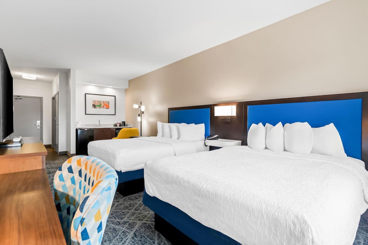 A hotel room with two beds, a desk, a chair, and a small kitchenette.