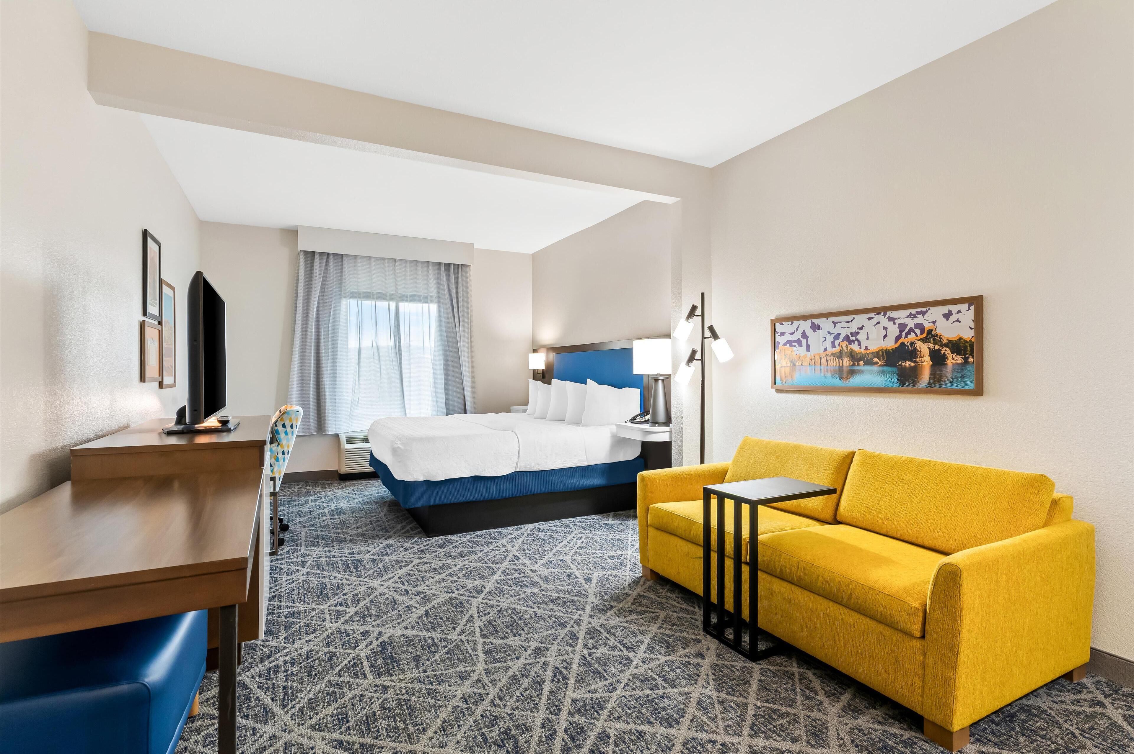 Quarto de hotel com cama, sofá amarelo, escrivaninha e um quadro na parede.