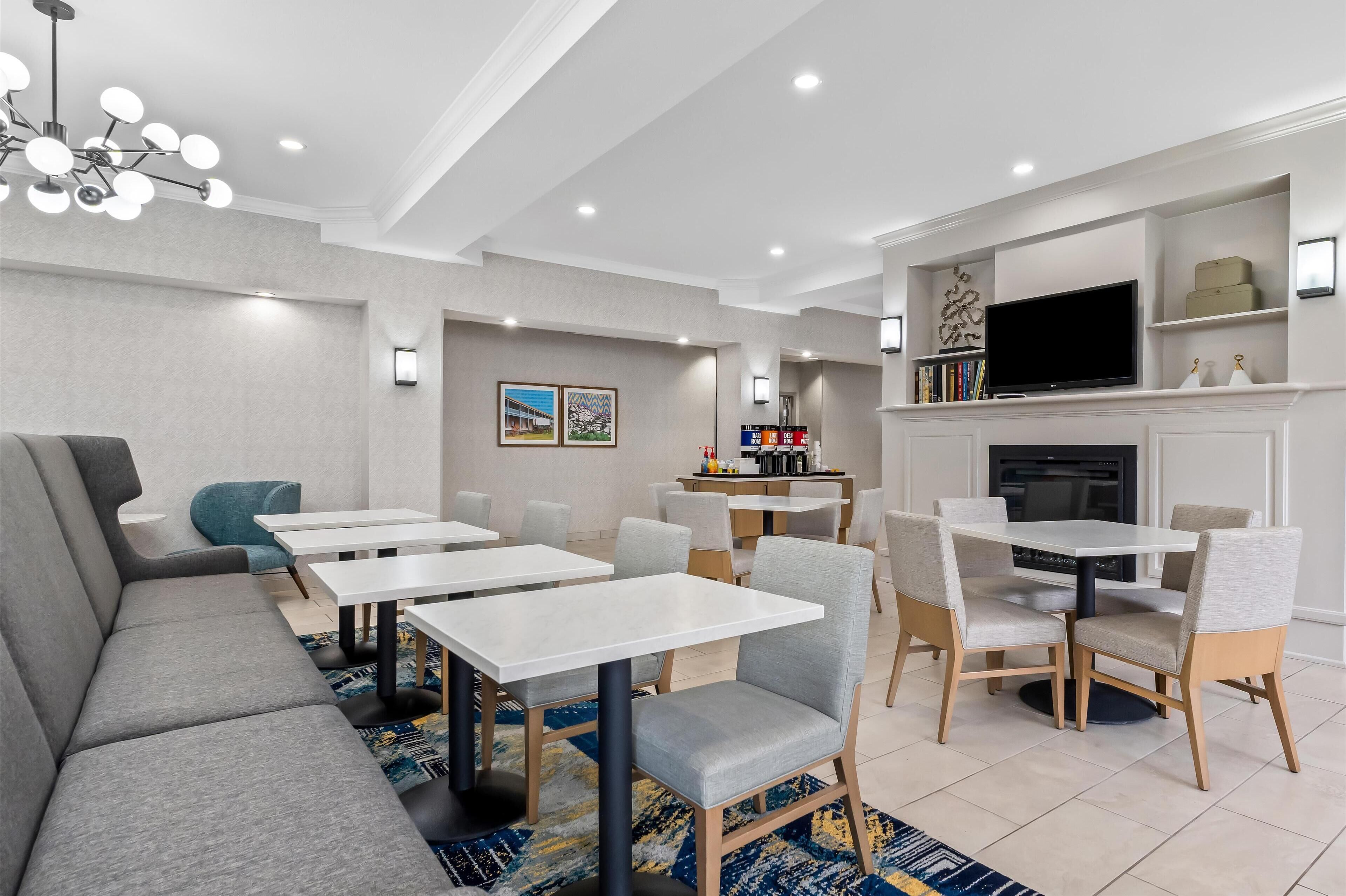 Uma sala de jantar moderna em hotel, com várias mesas, cadeiras, um sofá, uma lareira e uma televisão.
