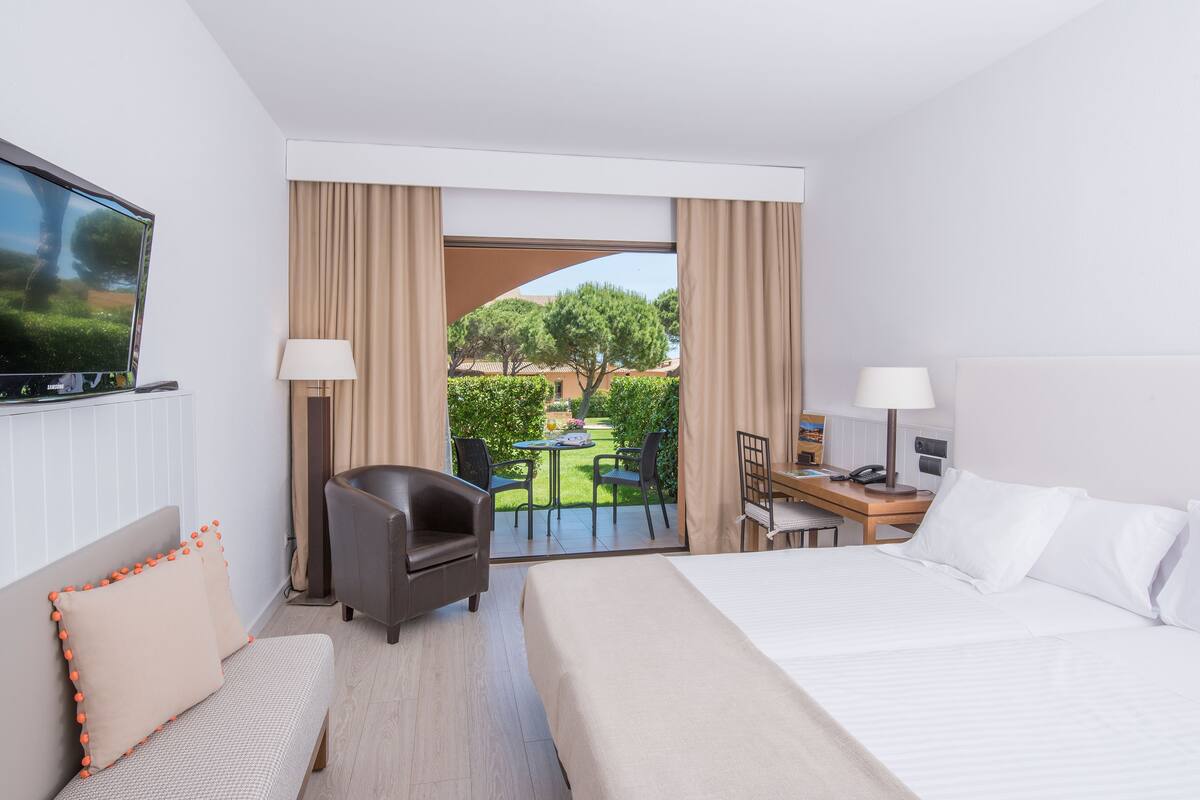 A hotel room with a bed, a desk, a chair, a TV, and a view of the garden.