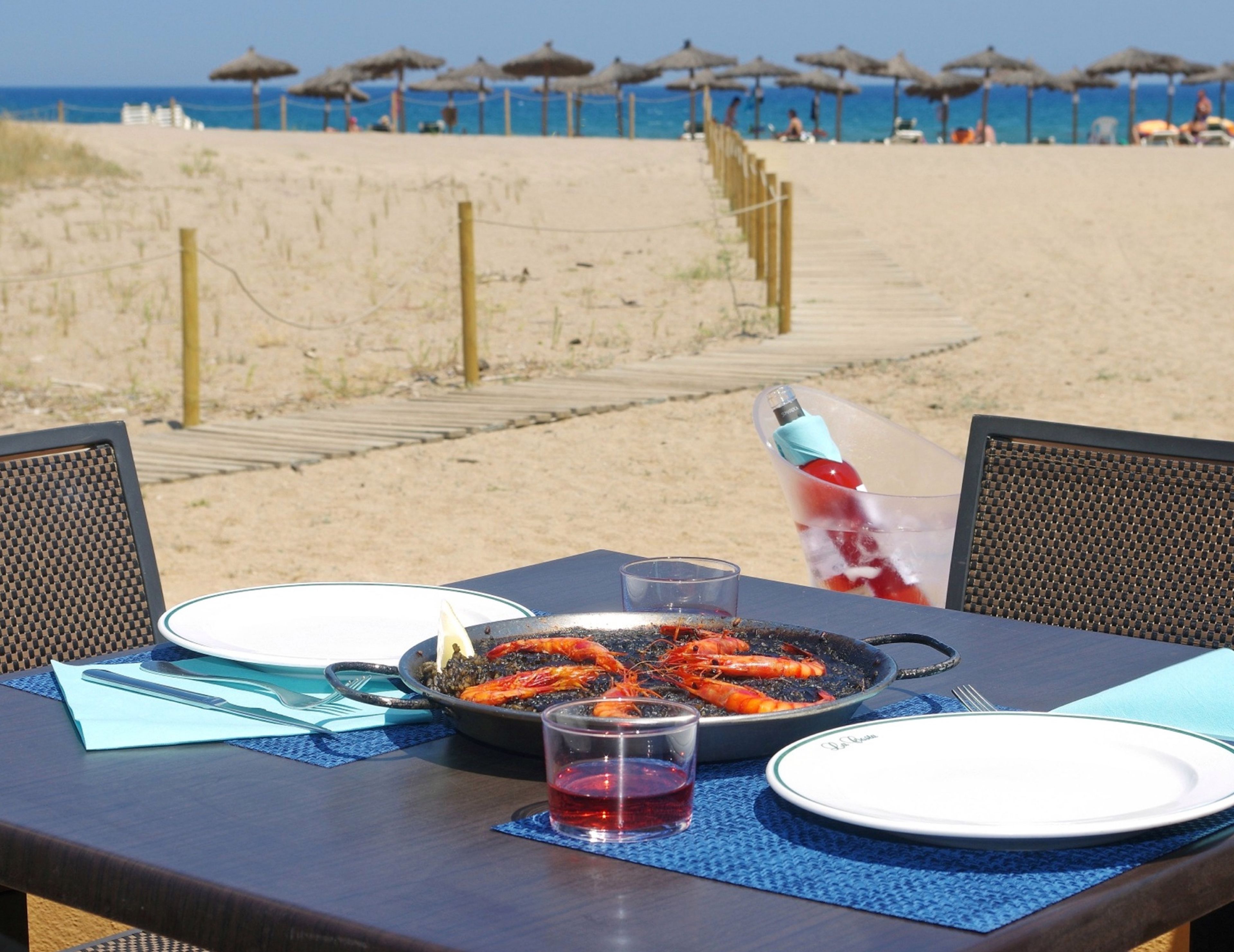 Mesa de comedor con mariscos, una copa de bebida roja y platos preparados para dos, con vista a una playa con sombrillas y un paseo de madera.