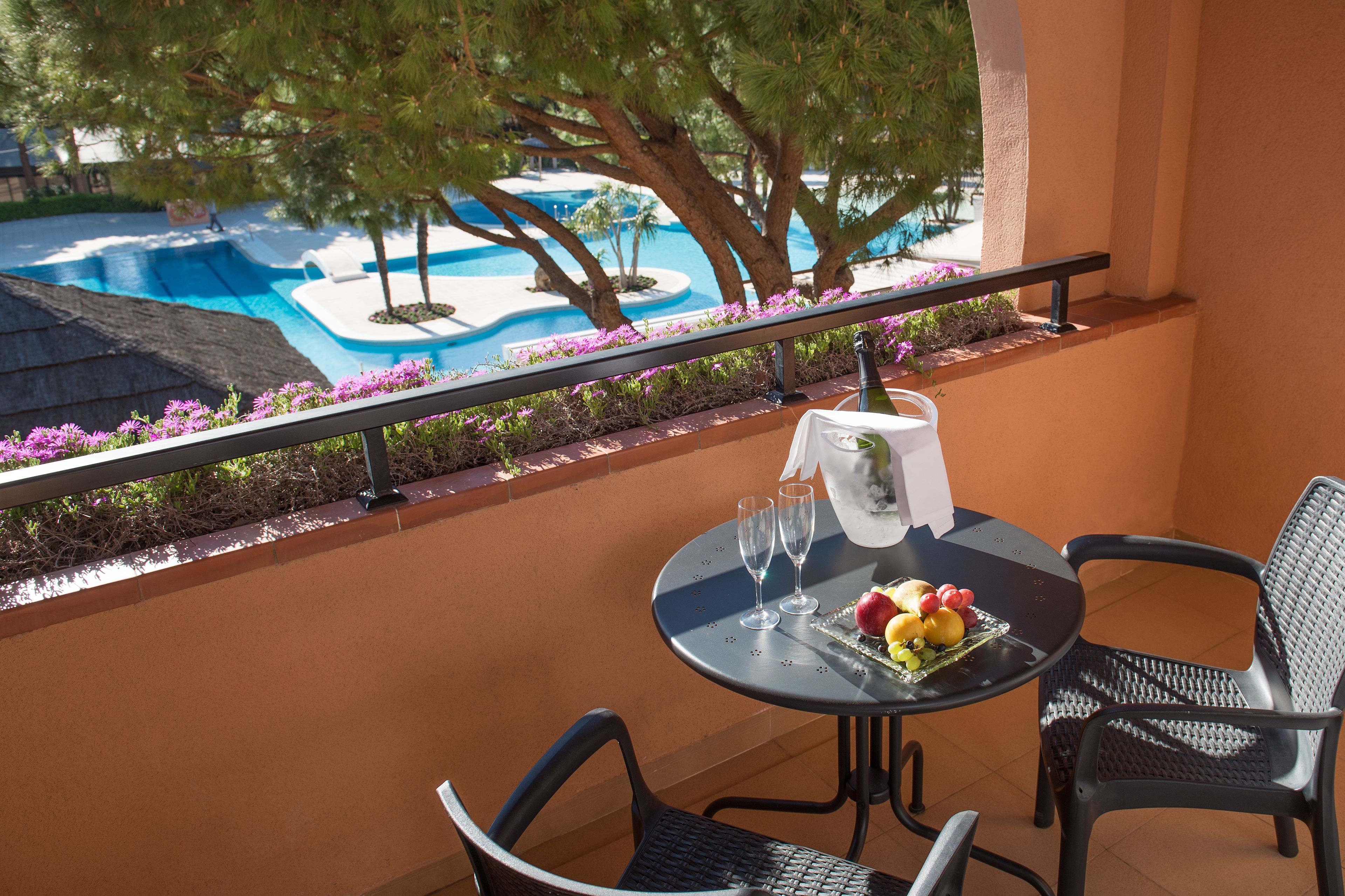 A balcony with a table set for two, overlooking a swimming pool and surrounded by flowers.